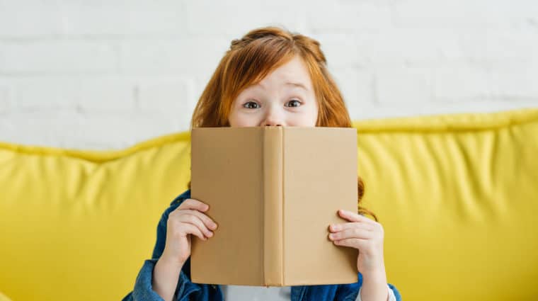 Child sitting on sofa and holding book in front of her face
